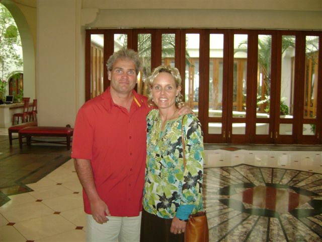 A man and a woman standing in a lobby. The man is wearing a red polo shirt and white pants while the woman has blonde hair. They are both smiling and looking at the camera. The lobby has a tiled floor with a circular pattern and there are large windows in the background that let in natural light. There are also some plants and furniture visible in the lobby.