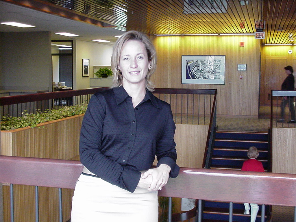 A woman standing in a lobby of a building. She is wearing a black blouse and a white skirt. She has blonde hair and is smiling at the camera. Behind her there is a staircase with a wooden railing and a small child sitting on the steps. On the wall there are several framed pictures and a sign that reads "Welcome to the lobby." The lobby has a high ceiling with wooden beams and a large window on the left side.