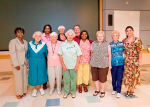 A group of nine women standing in a large room with a blue and white checkered floor. They are all smiling and appear to be posing for a group photo. The women are of different ages and ethnicities and they are all dressed in formal attire. The woman on the far left is wearing a blue dress with a white collar and a white blouse while the woman in the center is wearing green scrubs and a pink shirt. The other women are wearing colorful dresses with floral patterns. The background of the image is a white wall with a projector screen.