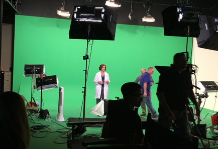 A green screen set up in a studio. There are several people in the image including a woman in a white lab coat standing in front of the screen. She appears to be giving a presentation or explaining something to the group. The studio is filled with various equipment and equipment including lights monitors and other equipment. The floor is covered in green carpet and there are several chairs and tables scattered around the room. The overall atmosphere of the image is professional and focused.