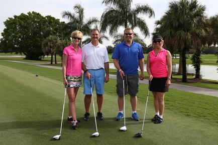 Four people standing on a golf course. They are all holding golf clubs and are posing for a photo. The person in the middle is wearing a white polo shirt and blue shorts while the person on the left is a blonde woman wearing a pink shirt and black shorts. The two people on the right are wearing pink shirts and black skirts. All four people are smiling and appear to be happy. In the background there are palm trees and a lake. The sky is blue and the weather appears to be sunny.