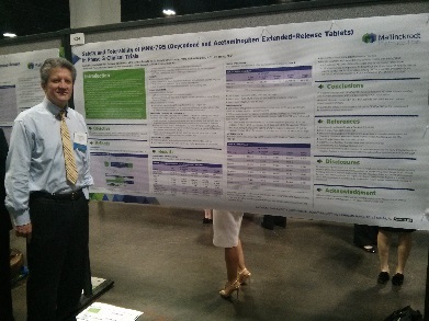 A man standing next to a large poster on a table. The poster is titled "Extended-Release Tablets" and appears to be a research paper. The man is wearing a blue shirt tie and glasses and is smiling at the camera. He is standing in front of the poster and is holding it up with both hands.<br /><br />The poster has multiple sections each with a title and a brief description of the topic. The title of the paper is written in bold letters at the top followed by headings for "Introduction" "Research" and "Introduction". Below the title there is a table with more information about the topic such as the date time and location. The table is set up in a conference room with other people in the background.