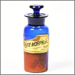 Of a small glass bottle with a blue body and a black lid. The bottle is cylindrical in shape and has a narrow neck. The body of the bottle is made of glass and is painted in a combination of blue and orange colors. On the front of the body there is a yellow label with black text that reads "Gran Morph.S.O.L." The label also has a small illustration of a mountain range in the background. The label appears to be old and worn with some areas of the glass appearing to be peeling off.