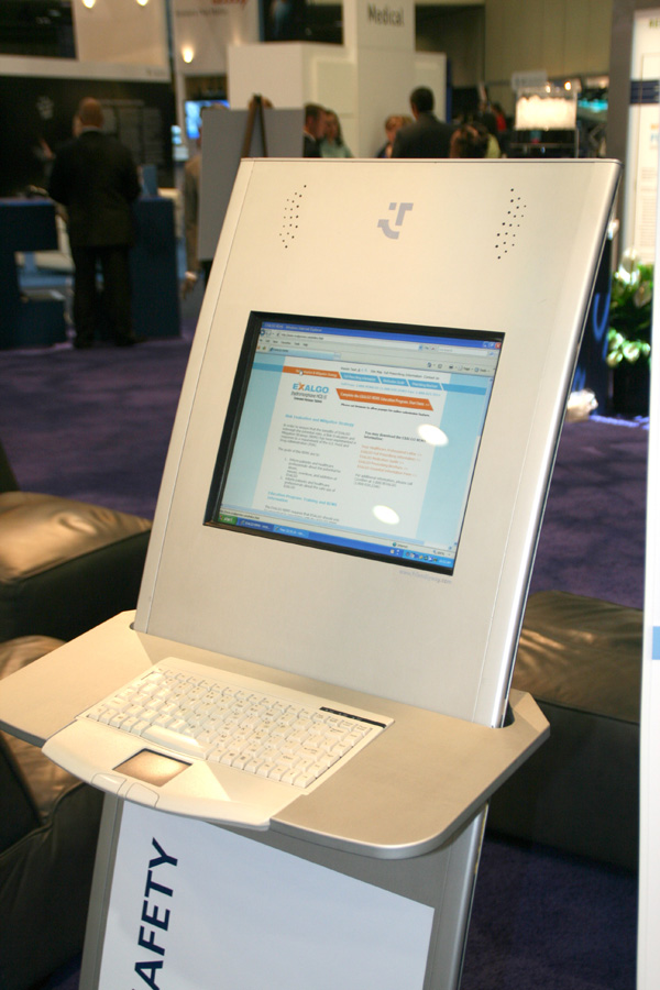 A white kiosk with a laptop on top of it. The kiosk is located in an exhibition hall with other booths and people in the background. The laptop screen displays a website with a blue and orange color scheme. The word "SAFETY" is written on the side of the kiosk in black letters.