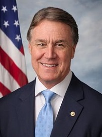 A professional headshot of a man in a suit and tie. He is standing in front of an American flag which is visible in the background. The man is smiling and looking directly at the camera. He has short dark hair and appears to be in his late 60s or early 70s. The suit is dark blue with a white shirt and a light blue tie. There is a small gold pin on his lapel. The background is a plain blue sky with white clouds.