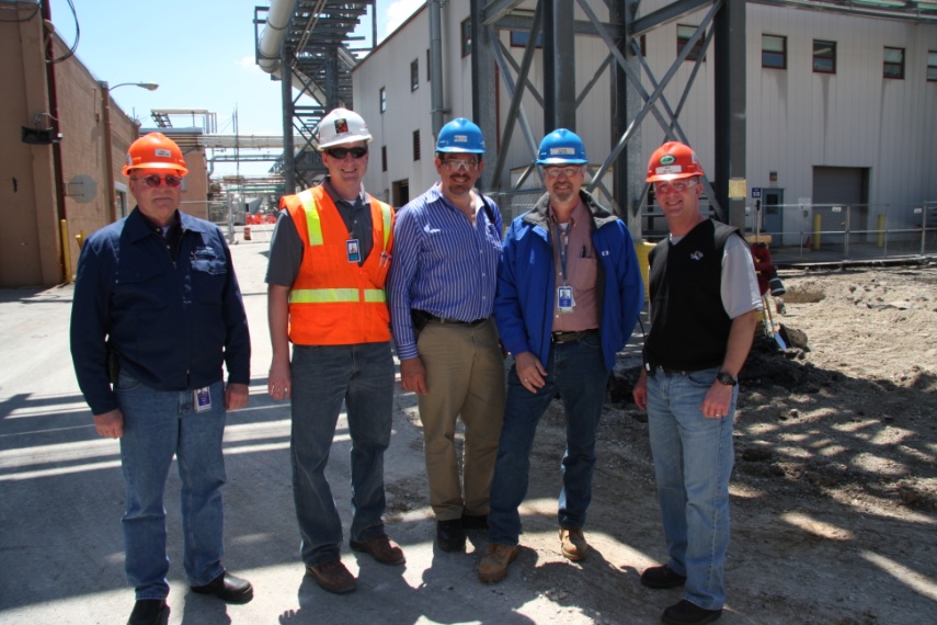 A group of five men standing in front of a construction site. They are all wearing hard hats and safety vests indicating that they are at a construction or industrial site. The men are standing close together and appear to be posing for a photo. In the background there is a large industrial building with a metal structure and a crane. The sky is blue and the ground is covered in dirt and debris. It appears to be a sunny day.