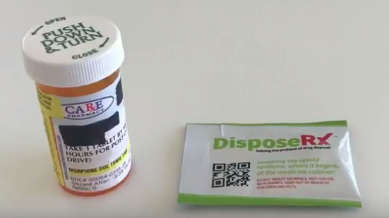 A small prescription bottle with a white cap and a green label. The label has the words "PUSH DOWN & TURN" written in bold black letters. Next to the bottle there is a small packet of Dispose Rx. The packet has a QR code on it. The background is white.