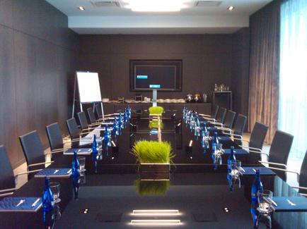 A large conference room with a long black table in the center. The table is covered with a black tablecloth and has several blue water bottles and glasses on it. There are several chairs around the table arranged in a semi-circle around it. On the right side of the image there is a large window with sheer curtains allowing natural light to enter the room. In the background there are two whiteboards and a flat-screen TV mounted on the wall. The room appears to be well-lit with recessed lighting.