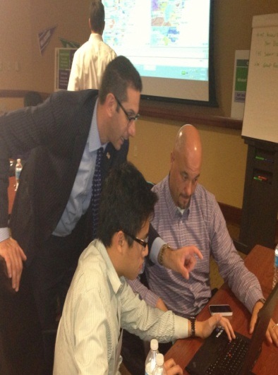 Three men in a meeting room. The man on the left is wearing a suit and tie and is leaning over a table looking at a laptop screen. He appears to be explaining something to the other two men who are sitting at the table and looking at the laptop screen with interest. The third man is also looking at his laptop screen and seems to be focused on the task at hand. There are water bottles and a mobile phone on the table. In the background there is a projector screen and a whiteboard on the wall.