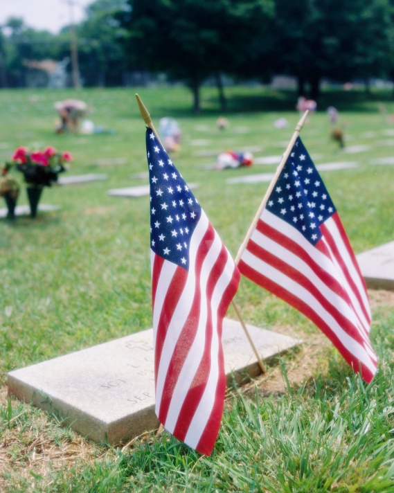 Two American flags on a grave in a cemetery. The flags are small and have a wooden stick attached to them. The flag on the left is slightly larger than the one on the right. The background shows a grassy field with other graves and flowers scattered around. The sky is overcast and there are trees in the distance. The overall mood of the image is somber and patriotic.