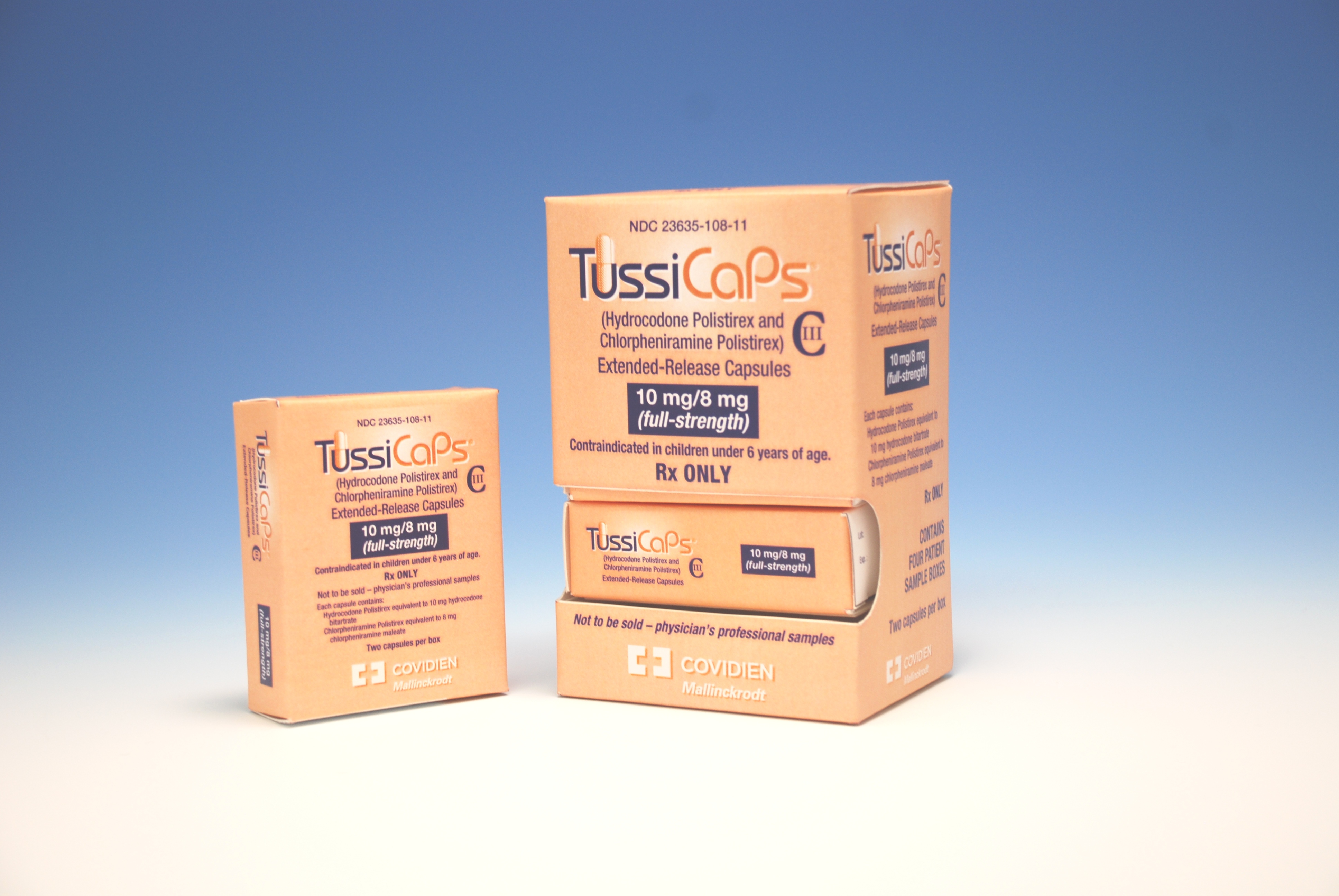 Two orange boxes of TussiCaps capsules. The boxes are rectangular in shape and have a white label on the front. The label has the brand name "TussiCaps" written in bold black letters at the top followed by the company name "Covidien" in smaller black letters. Below that there is a description of the product which states that it is a 10mg/ml capsule with a strength of 10mg. The box on the left is smaller and has the same label as the one on the right. The background is a light blue color.