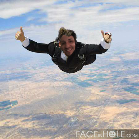 A young man skydiving in the sky with his arms stretched out wide. He is wearing a black suit and has a big smile on his face. The background shows a vast landscape of land and fields with a clear blue sky above. The man appears to be in mid-air with his eyes closed and his arms raised in a thumbs up gesture. The image appears to have been taken from a high vantage point looking down on the man and the landscape below.