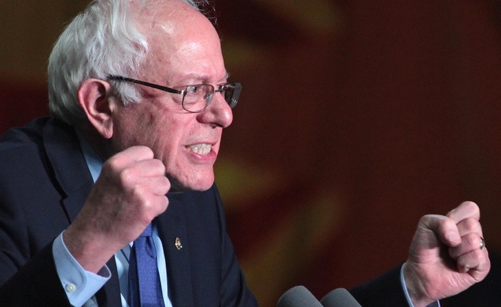 Bernie Sanders an American politician speaking at a podium with multiple microphones in front of him. He is wearing a dark suit and tie and has a serious expression on his face. His right hand is raised in a fist-like gesture as if he is making a point or making a statement. The background is blurred but it appears to be a stage with a red curtain. Sanders is likely giving a speech or addressing an audience.