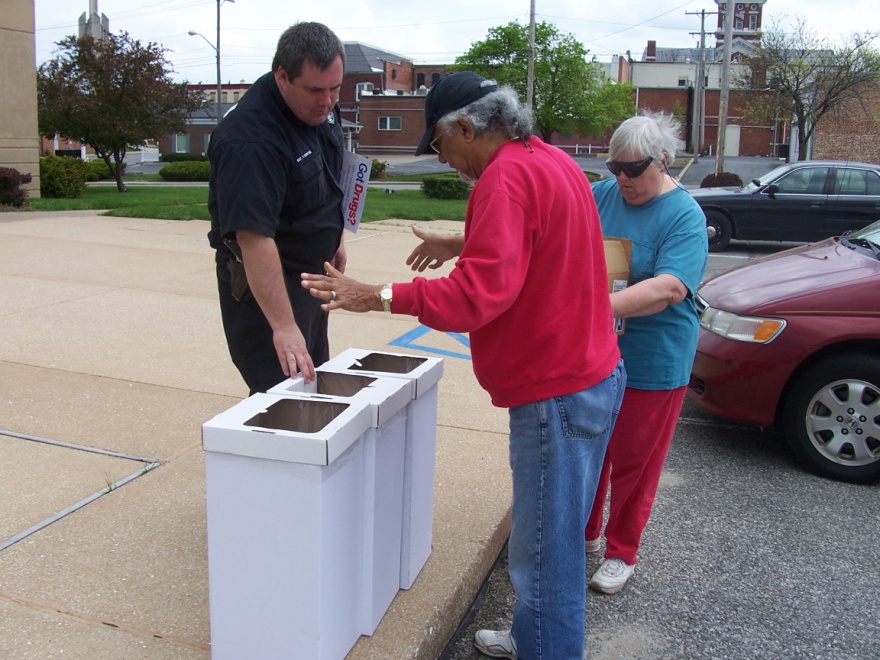 Three people standing on a sidewalk in a parking lot. On the left side of the image there is a man wearing a black jacket and a woman wearing a blue shirt. They are standing next to a white trash can. The man is pointing at the trash can with his hand while the woman is looking at it with a concerned expression. In the background there are several cars parked on the street and a building with a clock tower. The sky is overcast and the ground is wet suggesting that it has recently rained.