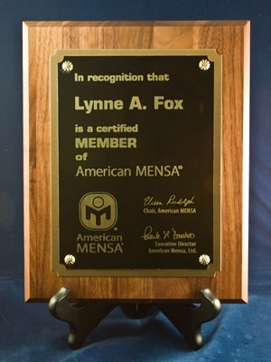 Of a wooden plaque with a black background. The plaque is rectangular in shape and has a gold border around the edges. On the front of the plaque there is a text that reads "In recognition that Lynne A. Fox is a certified member of American MENSA". Below the text there are two logos - one is the American Mensa logo and the other is the logo of the organization. The text is written in white and is in a serif font. There are also two small gold stars on either side of the text. The background is a solid blue color.