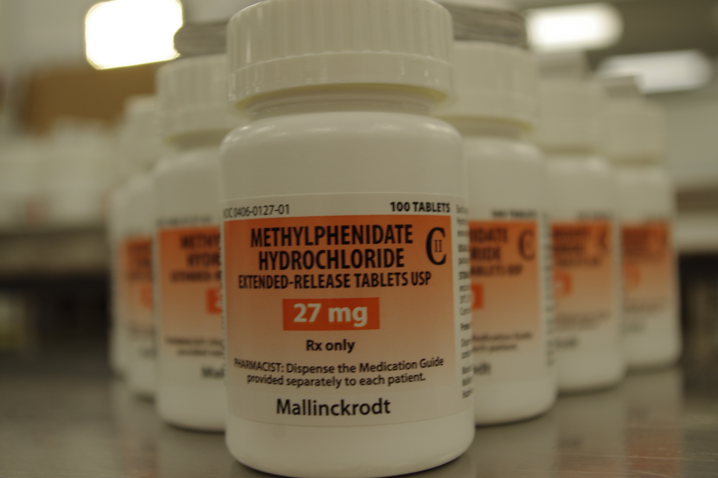 A row of white prescription bottles with orange labels. The bottles are arranged in a row on a metal shelf. The label on the bottles reads "Methylphenidate Hydrochloride Extended-Release Tablets USP 27 mg Rx only". The label also mentions that the bottles contain 100 tablets. The background is blurred but it appears to be a pharmacy or medical facility.