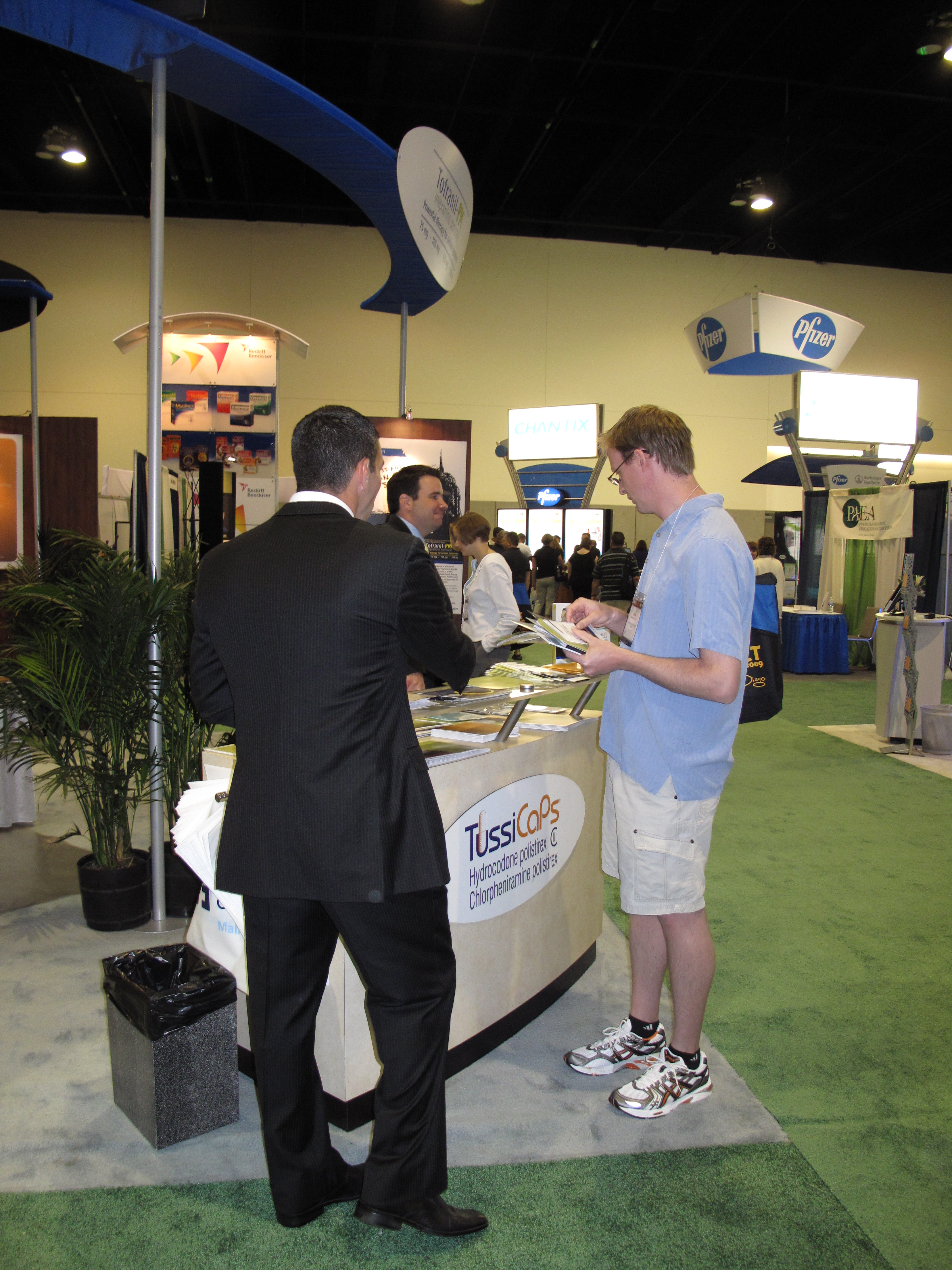 A trade show booth with a man in a black suit standing in front of it. He is holding a clipboard and appears to be explaining something to another man who is standing next to the booth. The booth has a white counter with a sign that reads "TussiCaps" and there are several other booths in the background. There are also several people in the booth some of whom are looking at the booth and others are engaged in conversation. The floor is covered in green carpet and there is a blue canopy on the left side of the image.