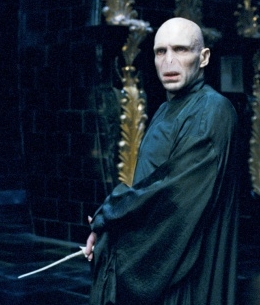 A man standing in a dimly lit room with a dark background. He is wearing a long black robe and is holding a sword in his right hand. The man appears to be in his late 40s or early 50s with a bald head and a serious expression on his face. He has a pale complexion and is looking directly at the camera. Behind him there are ornate gold pillars and a brick wall. The overall mood of the image is ominous and foreboding.