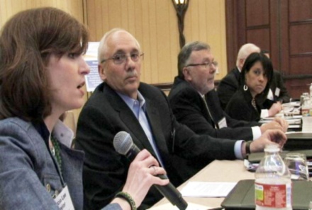 A group of people sitting at a long table in a conference room. There are six people in the image three men and two women all of whom appear to be engaged in a discussion. The woman on the left is holding a microphone and appears to be speaking while the man in the middle is listening attentively. The man on the right is wearing a suit and glasses and the woman next to him is also speaking into the microphone. The table is covered with a white tablecloth and there are water bottles and papers on it. The room has wooden paneling on the walls and a chandelier hanging from the ceiling.