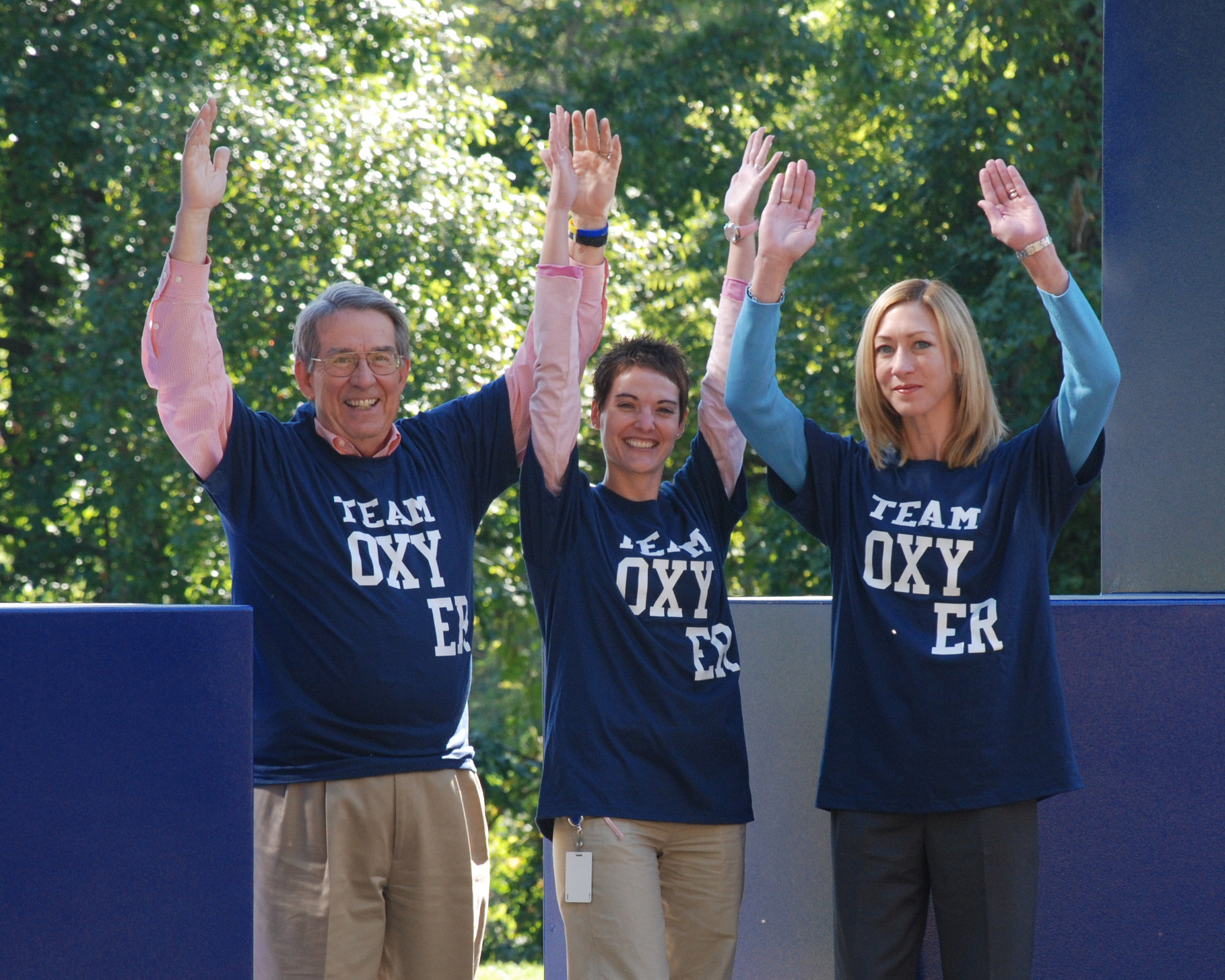 Three people two men and a woman standing on a stage with their hands raised in the air. They are all wearing blue t-shirts with the words "Team Oxygen" printed on them. The man on the left is an older man with gray hair and glasses the man in the middle is a young man with short dark hair and the woman on the right is a blonde woman with shoulder-length hair. All three are smiling and appear to be happy. The background is a blue wall with trees visible behind them.