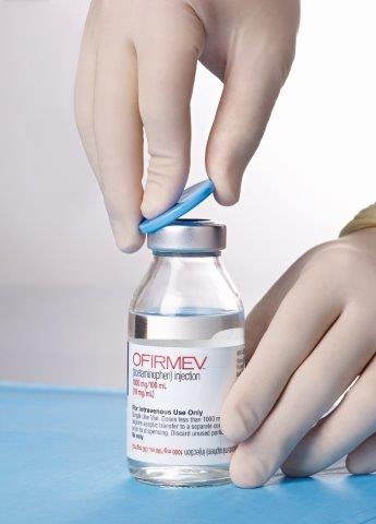 A hand wearing a white glove holding a small vial with a blue cap. The vial is filled with a clear liquid and has a label on it that reads "OFIRMEV". The background is a plain white surface. The hand is holding the vial and appears to be filling it with the liquid. The label also has some text on it but it is not clear what it says.