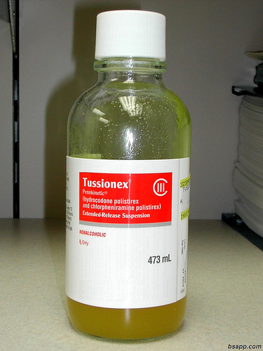 A bottle of Tussionex a type of liquid sitting on a countertop. The bottle is made of clear glass and has a white cap. The label on the bottle is red and white with the brand name "Tassoni" written in bold black letters. Below the label there is a description of the product which states that it is an extended-release suspension of 473 ml. The liquid inside the bottle appears to be a yellowish-orange color. The background is blurred but it seems to be an office or laboratory setting.