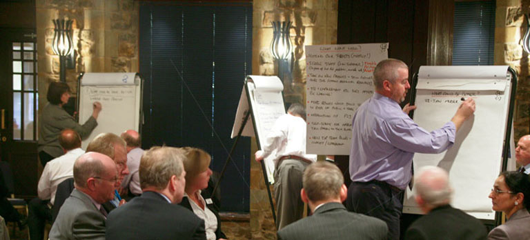 A group of people gathered in a room with stone walls and chandeliers. There are two whiteboards in the center of the room one on the left side of the image and the other on the right side. The man in the purple shirt is standing in front of one of the whiteboards and is writing on it with a marker. He appears to be giving a presentation or presentation to the group. The people in the room are sitting in chairs and are attentively listening to the man. They are all dressed in formal business attire and appear to be engaged in the presentation.