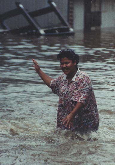 A woman standing in a flooded street. She is wearing a red and white patterned shirt and has her arms stretched out to the side. The water is up to her knees. In the background there is a building that is partially submerged in the water.