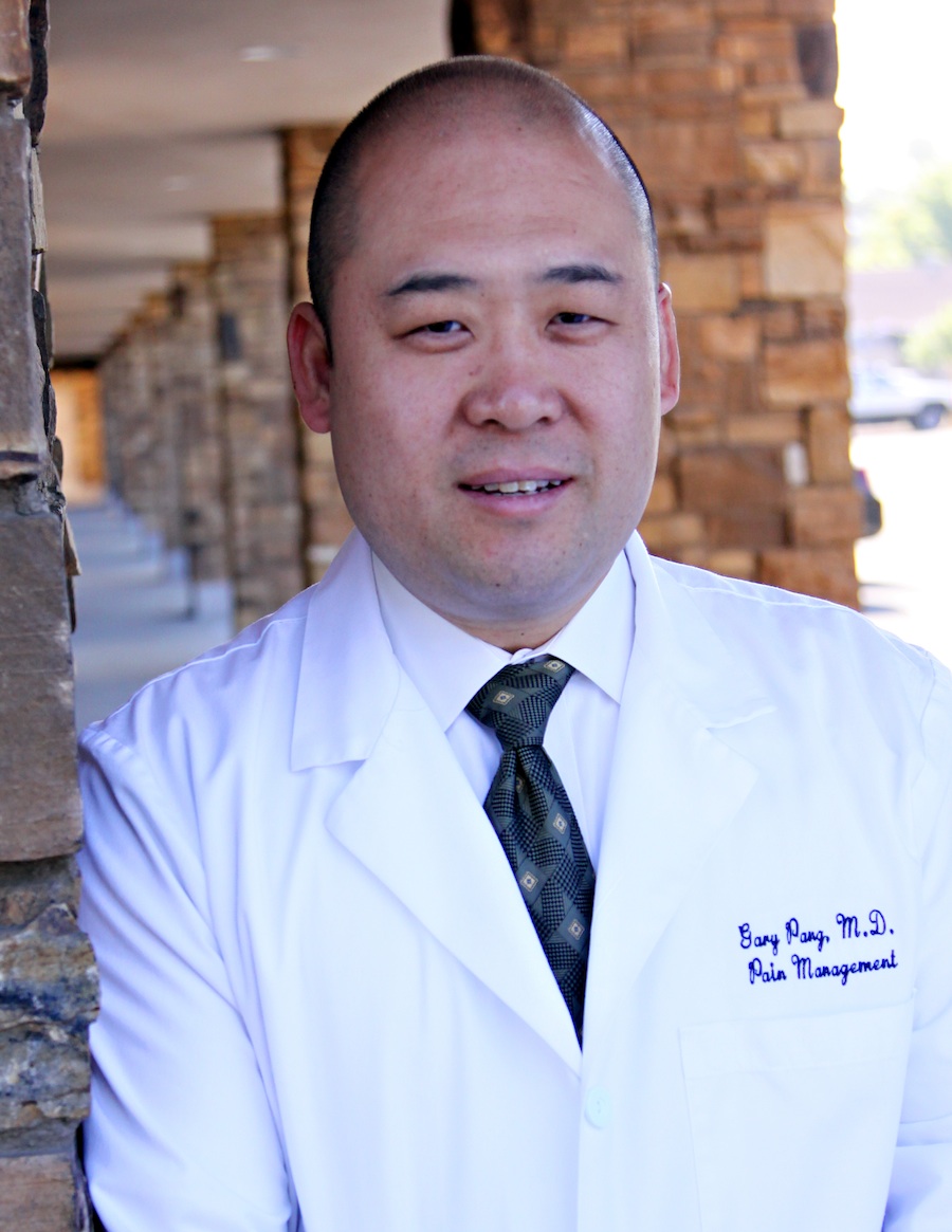 A portrait of a middle-aged Asian man wearing a white lab coat and a blue tie. He is standing in front of a stone wall with a blurred background. The man is smiling and looking directly at the camera. On the right side of his coat there is a name tag that reads "Barry Pang MD Pain Management." He appears to be in his late 40s or early 50s.