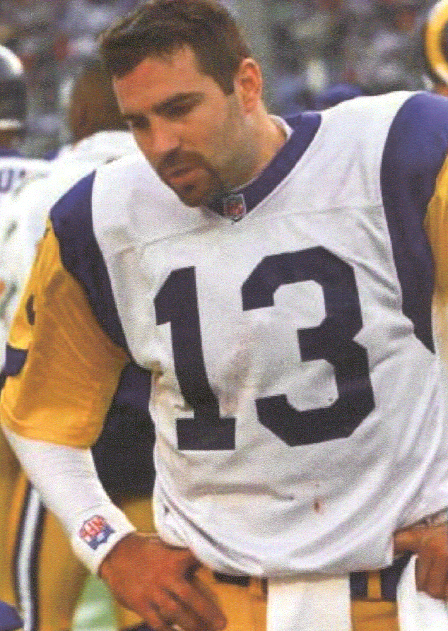 A man wearing a white and blue football jersey with the number 13 on it. He is standing on a football field with other players in the background. The man appears to be in his late twenties or early thirties and has short dark hair and a beard. He has a serious expression on his face and is looking off to the side. The jersey has the NFL logo on the left side of his chest and the number "13" on the right side.