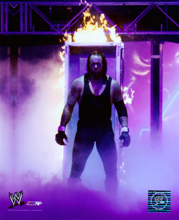 A photograph of a professional wrestler The Undertaker standing in front of a large fire pit. He is wearing a black tank top and black pants and has long dark hair and a beard. His arms are crossed over his chest and he is looking off to the side with a serious expression on his face. The background is dark with purple and blue lights shining down on the wrestler. The WWE logo is visible in the bottom right corner of the image.