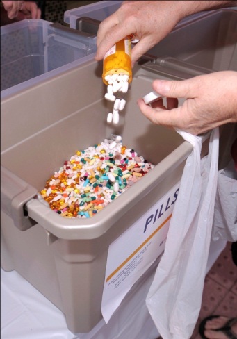 A person's hand holding an orange prescription bottle and dropping it into a beige plastic bin filled with colorful pills. The bin has a label on it that reads "PILLS". The person is holding a white plastic bag over the bin and appears to be in the process of dropping the pills into it. The background is blurred but it seems like the person is standing in a room with other people in the background.