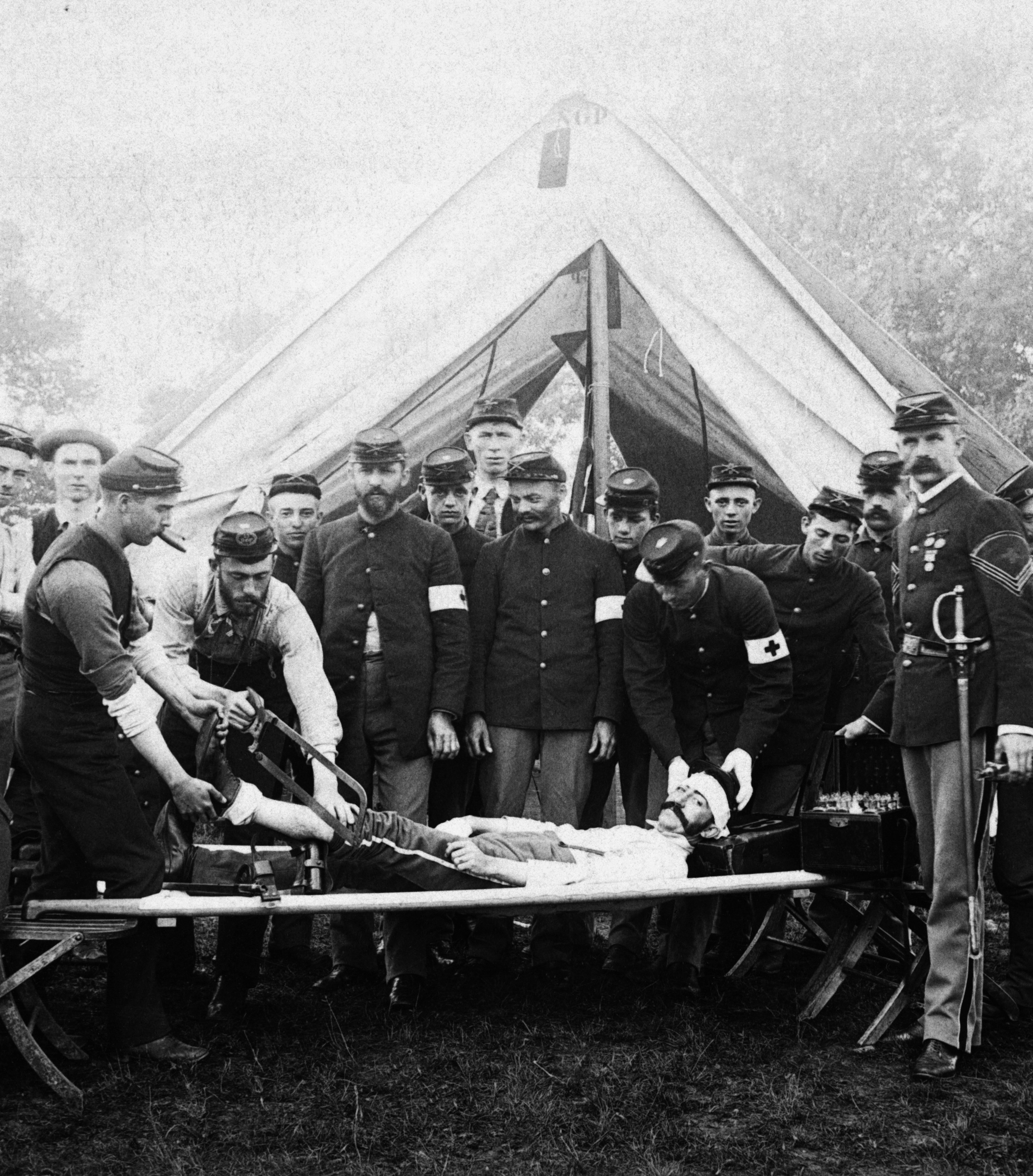 A black and white photograph of a group of men in military uniforms gathered around a man lying on a stretcher. The man appears to be injured or dead as he is lying on his back with his head resting on the stretcher and his arms stretched out to the sides. The men are standing around him some of them are holding medical equipment while others are looking at him attentively. In the background there is a tent and trees suggesting that the photo was taken in a rural area. The overall mood of the image is somber.