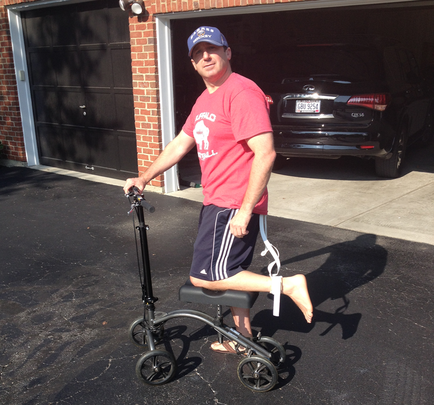 A man standing in front of a garage with a black car parked in the background. He is wearing a red t-shirt blue shorts and a baseball cap. He has a prosthetic leg that is attached to a walker with a white strap. The man is holding onto the handlebars of the walker and appears to be in the process of walking. The walker has four wheels and a seat for the man to sit on. The garage has a brick wall and a garage door.