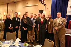 A group of nine people standing in a conference room. They are all dressed in formal attire and are posing for a photo. The room has a blue curtain on the right side and a banner on the left side with the logo of a company. There are tables and chairs in the background and a few people can be seen sitting at the tables. The people in the photo appear to be of different ages and genders and they are all smiling and looking at the camera. It seems like they are at an event or gathering.