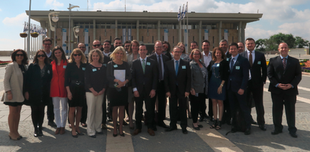 A group of people standing in front of a large building with a blue sky and white clouds in the background. The building appears to be a government building with multiple levels and a flagpole with the Israeli flag flying in front. There are around 20 people in the group all dressed in formal business attire and smiling at the camera. They are standing in a line facing the camera and appear to be posing for a group photo. The people are of different ages genders and ethnicity's. Some of them are wearing suits and ties while others are wearing casual clothes. The group is standing on a tiled surface and there are trees and bushes on either side of the building.