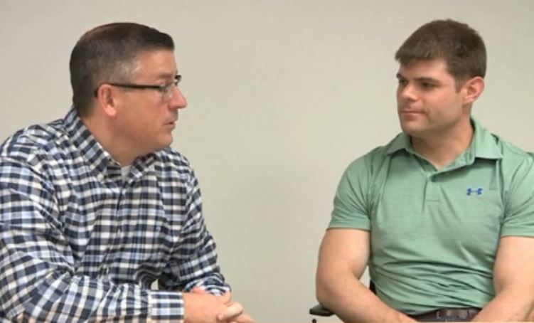 Two men sitting in a meeting room engaged in a conversation. The man on the left is wearing a blue and white checkered shirt and glasses and he appears to be in his late 40s or early 50s. He has short dark hair and is looking off to the side with a serious expression on his face. On the right is a young man wearing a green polo shirt with a blue Under Armour logo on it. He is sitting in front of a whiteboard and is listening intently to the other man. Both men are sitting on chairs and appear to be engaged in the conversation.