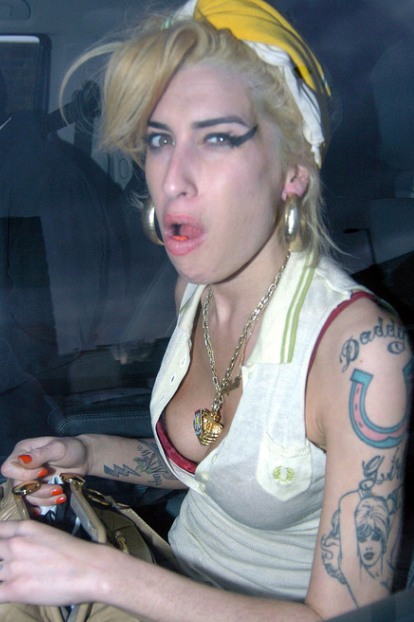 A young woman sitting in the back seat of a car. She is wearing a white sleeveless top with a yellow headband tied around her head. She has blonde hair styled in loose waves and is wearing large hoop earrings and a gold necklace with a pendant. She also has multiple tattoos on her arms and chest. The woman is holding a purse in her left hand and appears to be looking at the camera with a surprised or shocked expression on her face. The background is blurred but it seems like she is inside the car.