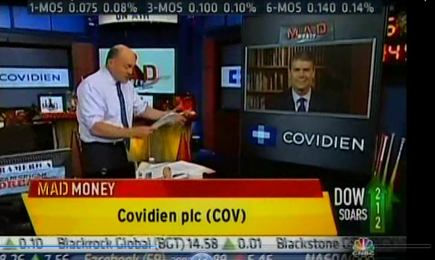 A screenshot of a news segment on a TV screen. The main focus of the image is on a man in a white shirt and tie who appears to be a news anchor. He is standing in front of a desk with a computer monitor and other news equipment in the background. The man is holding a piece of paper in his hands and is looking at it intently. On the right side of the screen there is a picture of a man wearing a suit and tie. The text on the screen reads "Covidien plc (COVID)". Below the man there are numbers and text that appear to be related to the news segment.