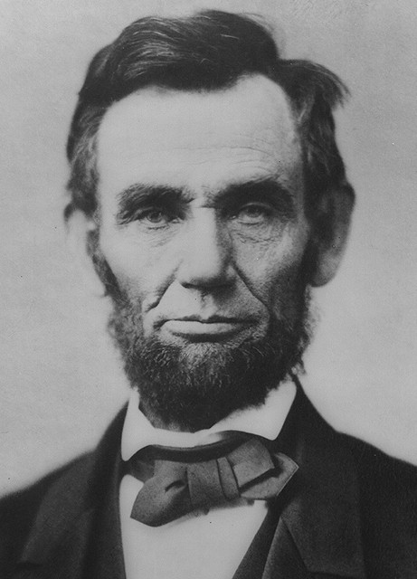 A black and white portrait of Abraham Lincoln the 16th President of the United States. He is shown from the waist up looking directly at the camera with a serious expression on his face. He has a full beard and mustache and his hair is neatly combed back. He appears to be wearing a suit and bowtie. The background is plain and blurred putting the focus on Lincoln's face.