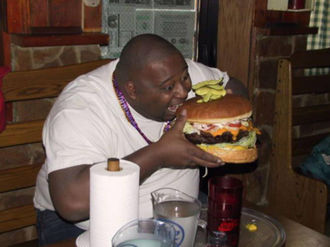 A man sitting at a wooden table in a restaurant. He is wearing a white t-shirt and a purple beaded necklace. He has a big burger in front of him with lettuce tomato cheese and pickles on top. The man is holding the burger with both hands and is taking a bite out of it. There are two glasses of water on the table next to him. The background shows a brick wall and a window.