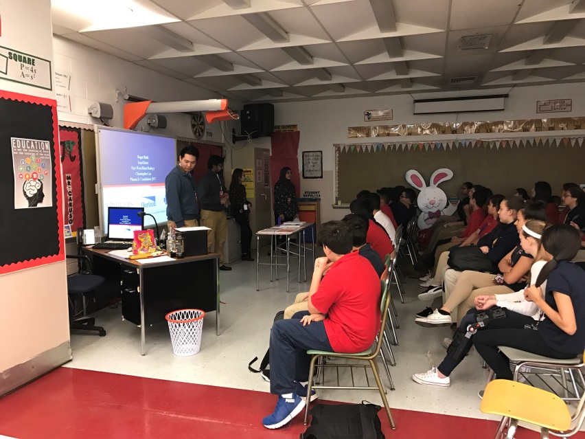 A classroom with a group of students sitting on chairs facing a projector screen. The students are attentively listening to a presentation given by a man standing at the front of the room. The room has a red carpet on the floor and a whiteboard on the left side of the image. There is a banner with a bunny design on the wall in the background. The classroom appears to be well-lit with natural light coming in from the windows.