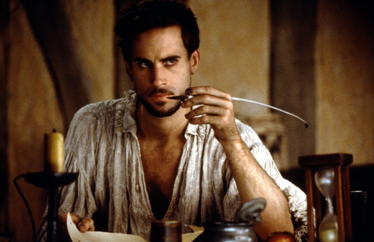 A man sitting at a table with a plate of food in front of him. He is wearing a white shirt and has a serious expression on his face. The man is holding a spoon in his right hand and appears to be deep in thought. On the table there is a candle with a flame a jar and an hourglass. The background is blurred but it seems like the man is in a dimly lit room with a wooden wall. The overall mood of the image is somber and contemplative.