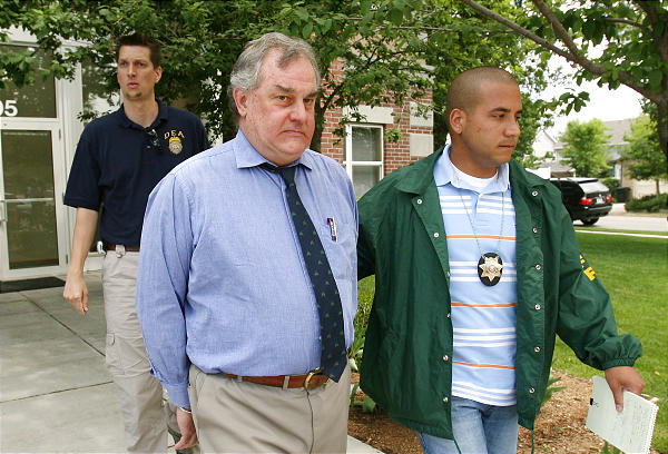 Three men walking on a sidewalk in front of a building. The man in the middle is an older man with gray hair and a blue shirt and tie. He appears to be in his late 60s or early 70s with a serious expression on his face. He is wearing a green jacket with a badge on the left side of his chest and a white shirt with a blue and orange striped shirt underneath. On the right side of the image there is a young man wearing a blue jacket with an orange and white striped shirt and a badge around his neck. He has short dark hair and is holding a piece of paper in his hand. Behind him there are two other men one wearing a navy blue polo shirt and khaki pants who appear to be police officers. The building in the background has a glass door and there are trees and bushes on the sidewalk.