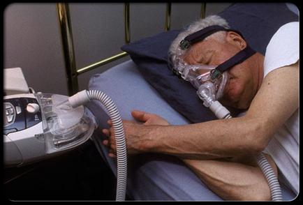 An elderly man sleeping in a hospital bed with an oxygen mask on. He is lying on his side with his head resting on a blue pillow and his eyes closed. The oxygen mask is connected to a white tube that is plugged into a medical device on the left side of the bed. The device appears to be a crap machine which is used to provide oxygen to the man's head and neck. The man is wearing a white t-shirt and is resting his head on the pillow. The background is a gray wall with a metal frame.