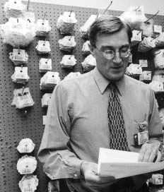 A black and white photograph of a man standing in front of a wall covered in small white objects. The man is wearing a collared shirt a tie and glasses. He is holding a piece of paper in his hands and appears to be looking at it intently. The objects on the wall are arranged in a grid-like pattern and are of different sizes and shapes. The background is plain and the focus is on the man and the objects.