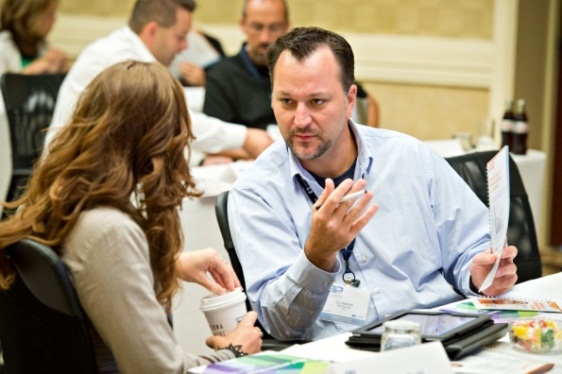 A man and a woman sitting at a table in a conference room. The man is wearing a blue collared shirt and has a name tag around his neck. He is holding a piece of paper in his hand and appears to be explaining something to the woman. The woman is sitting next to him and is looking at him attentively. There are other people in the background but they are not visible. The table is covered with a white tablecloth and there are various items on it including a laptop a cup of coffee and some papers. It appears that the man and woman are engaged in a discussion or brainstorming session.