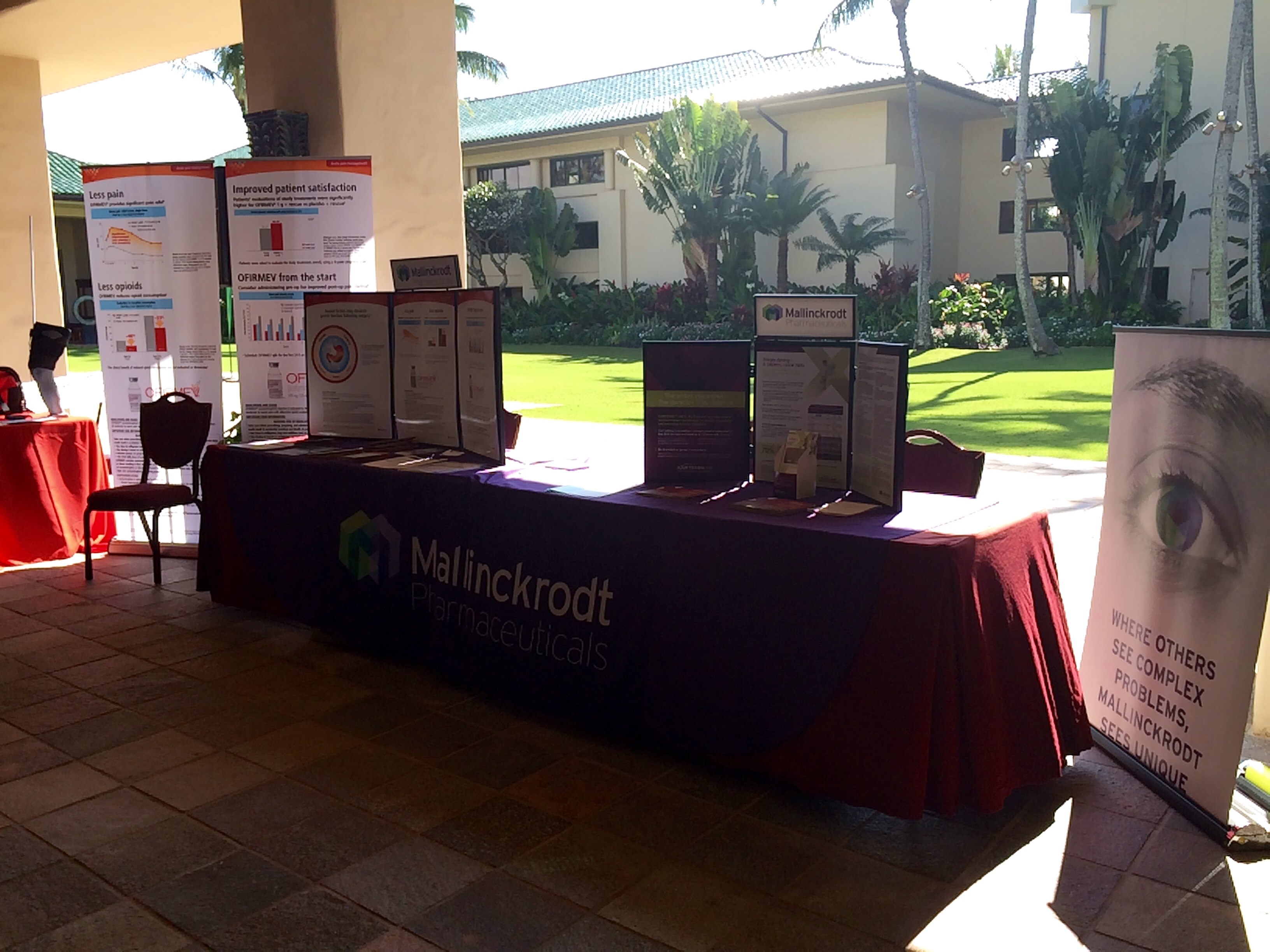 A table set up in a courtyard with a red tablecloth. On the table there are several posters and brochures with information about the company's products and services. The table is covered with a black tablecloth and there is a banner on the right side of the image that reads "Mallinckrodt Pharmaceuticals". In the background there is an outdoor area with palm trees and a building. The sky is blue and the sun is shining creating a warm and inviting atmosphere.