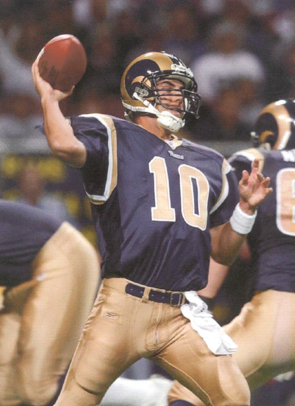 A football player in action during a game. He is wearing a navy blue jersey with the number 10 on it and a gold helmet with the team's logo on it. The player is holding the football in his right hand and appears to be in the process of throwing it. In the background there are other players from both teams wearing navy blue jerseys and gold helmets. The stands are filled with spectators and the field is covered in green turf. The image appears to have been taken during the day.