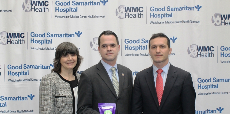 Three people standing in front of a backdrop with the logos of Good Samaritan Hospital and WMC Health. The person in the middle is holding a purple award which appears to be an award for a medical treatment. On the left is a woman with shoulder-length dark hair and bangs wearing a black and white checkered blazer. Next to her is a man in a suit and tie and on the right is a young man with short dark hair. All three people are smiling and appear to be posing for the photo.